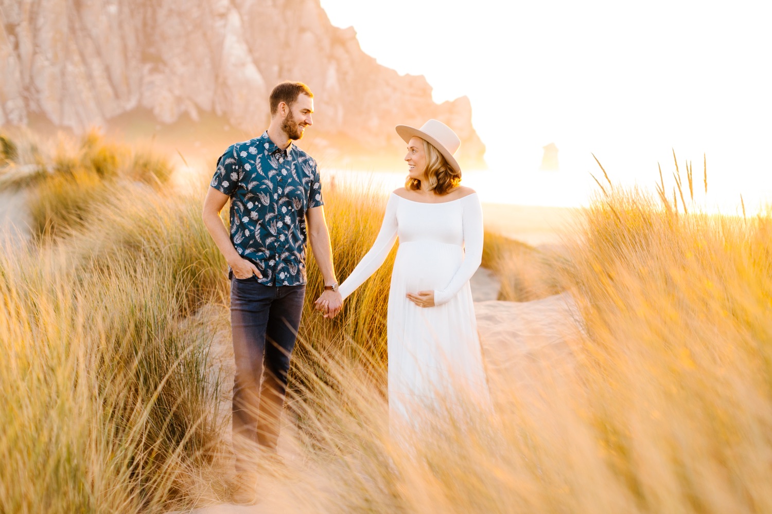 Couple holding hands while in the dunes at Morro bay beach with morro rock in the background at maternity session with Tayler Enerle