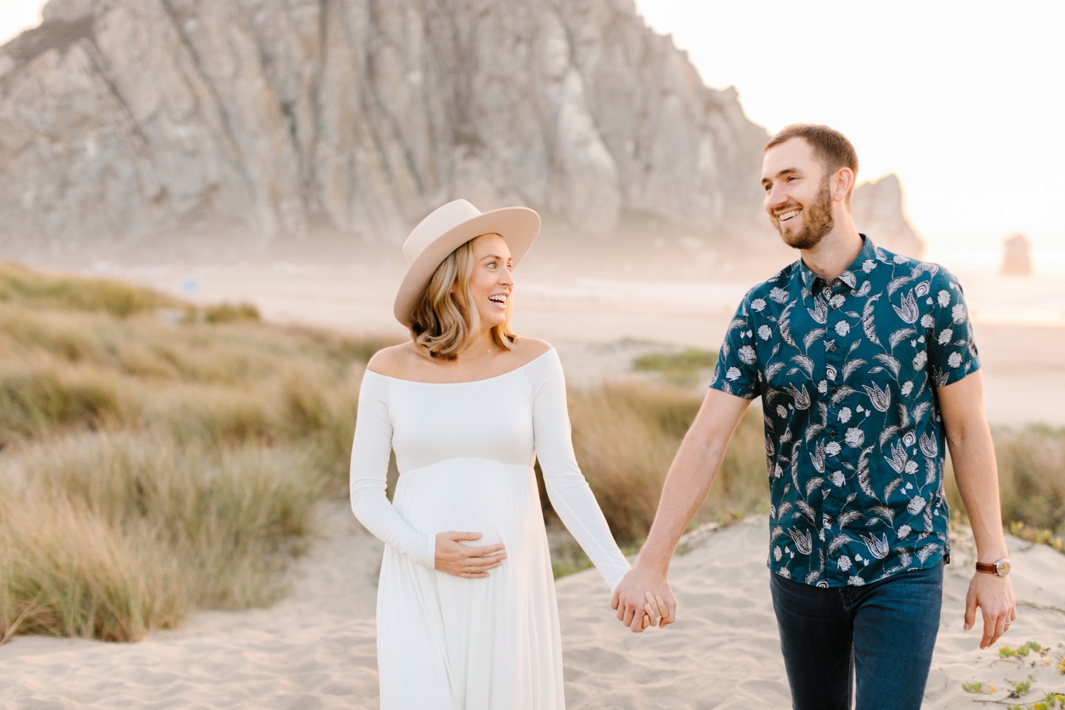 couple walking hand in hand on the sand dunes for their beach maternity session at Morro Bay rock, california. Woman is wearing a white dress and light colored hat, the man is wearing a blue and white patterned button up shirt and they are smiling at eachother during session photographed by newborn photographer, Tayler Enerle