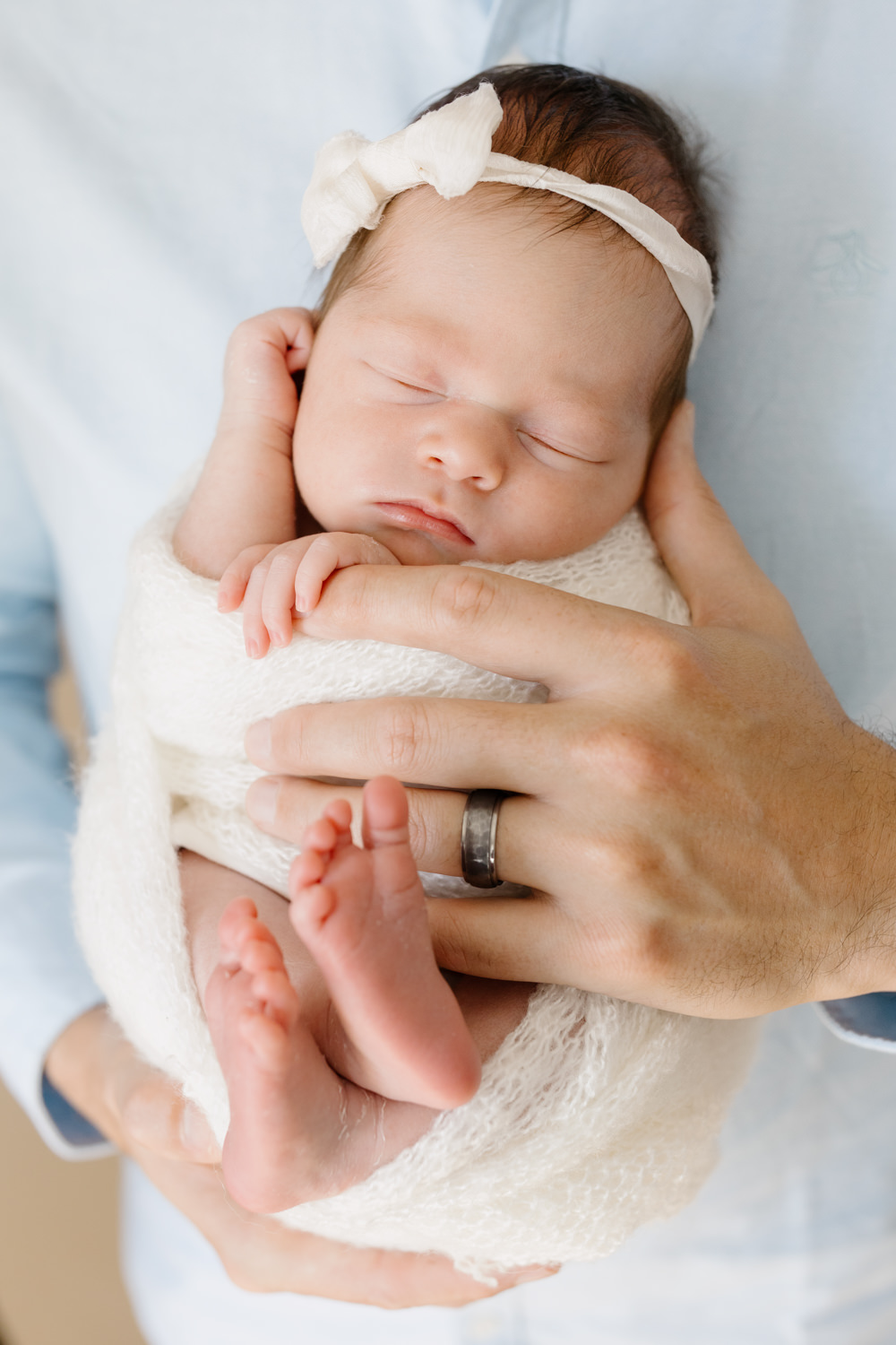 A portrait of a newborn baby girl held by her father, the image is a close up of her father's hands and her swaddled in a white wrap with a white bow on her head, photos taken at Tayler Enerle's Los Osos Newborn portrait studio