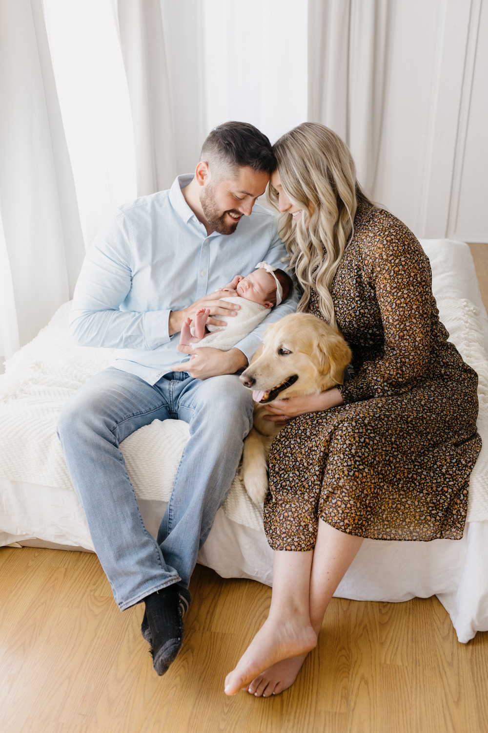 Los Osos Newborn photography session with golden retriever, parents sitting on bed holding their newborn baby who is swaddled in a soft white wrap with a white bow. They are looking at her adoringly, with their golden retriever dog laying cuddled up between them all. 