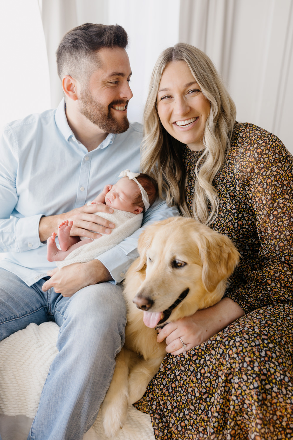 A photo from a newborn session with golden retriever including A smiling man and woman holding their newborn daughter, they are sitting on a white bed with their golden retriever between them at tayler Enerle's newborn photographer studio in Los Osos, the man is looking at his wife adoringly. 