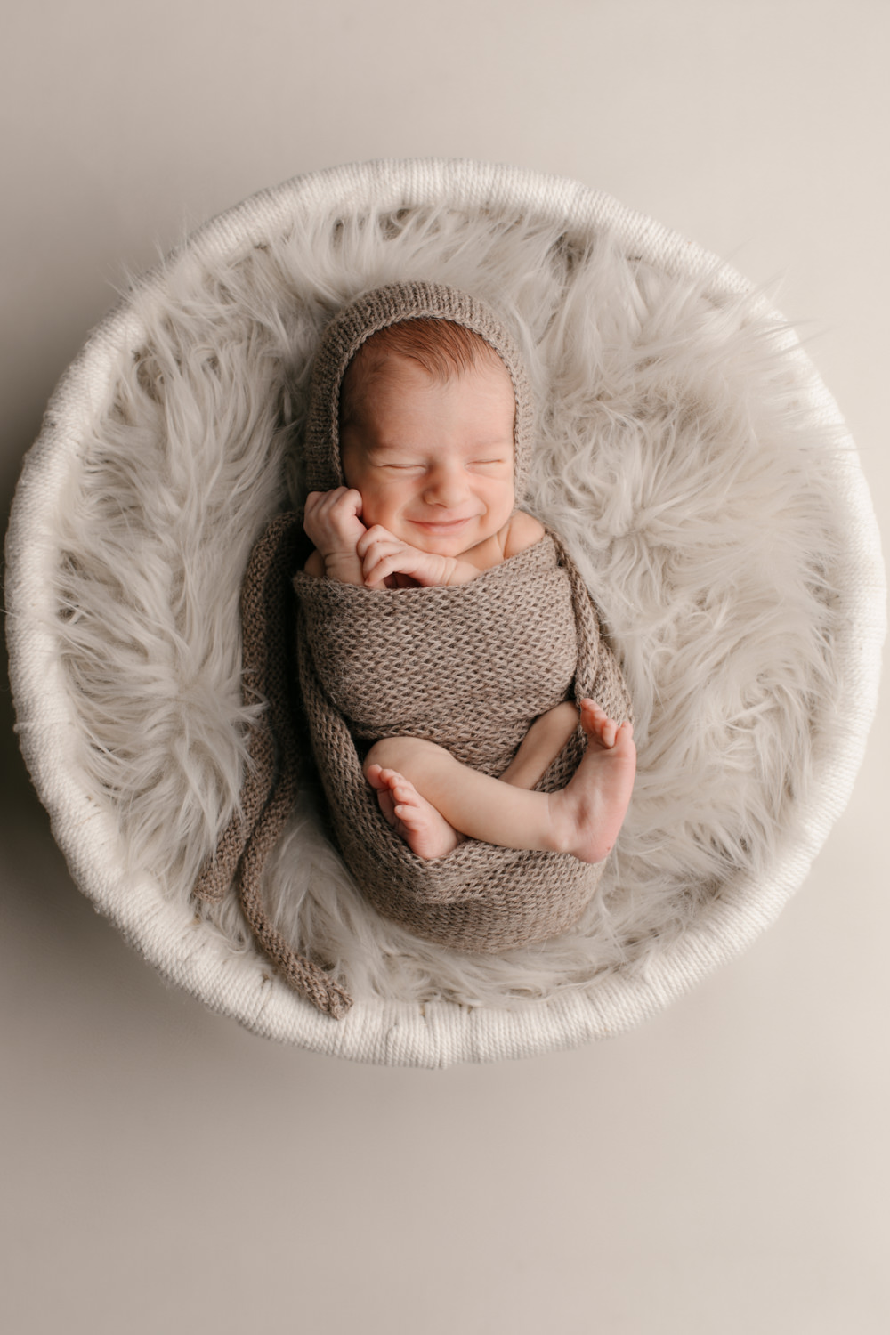 a newborn baby swaddled in a tan wrap with his feet and hands showing, he is laid on a fur lined basket, the baby is smiling in the basket which is in Tayler Enerle's San Luis Obispo County newborn photographer's studio in Los Osos California