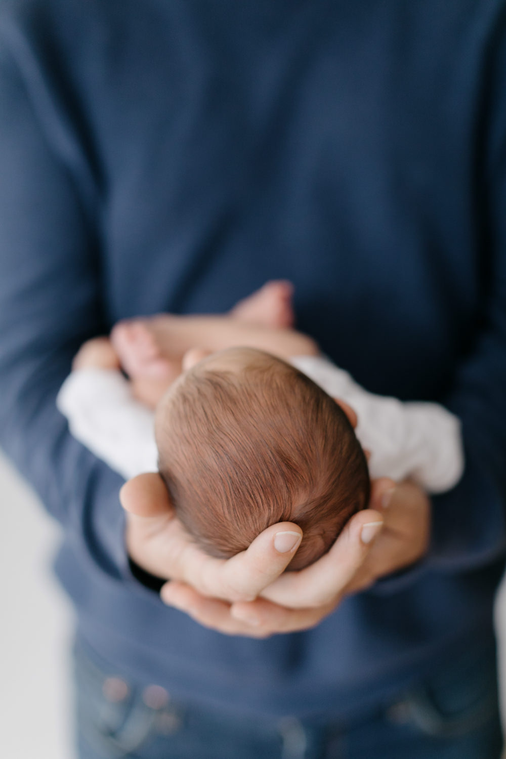 A close up portrait of a father's hands holding his small newborn baby, only the father's hands and the baby's hair and the out of focus rest of the body of the baby showing in a newborn portrait taken by San Luis Obispo newborn photographer, Tayler Enerle.