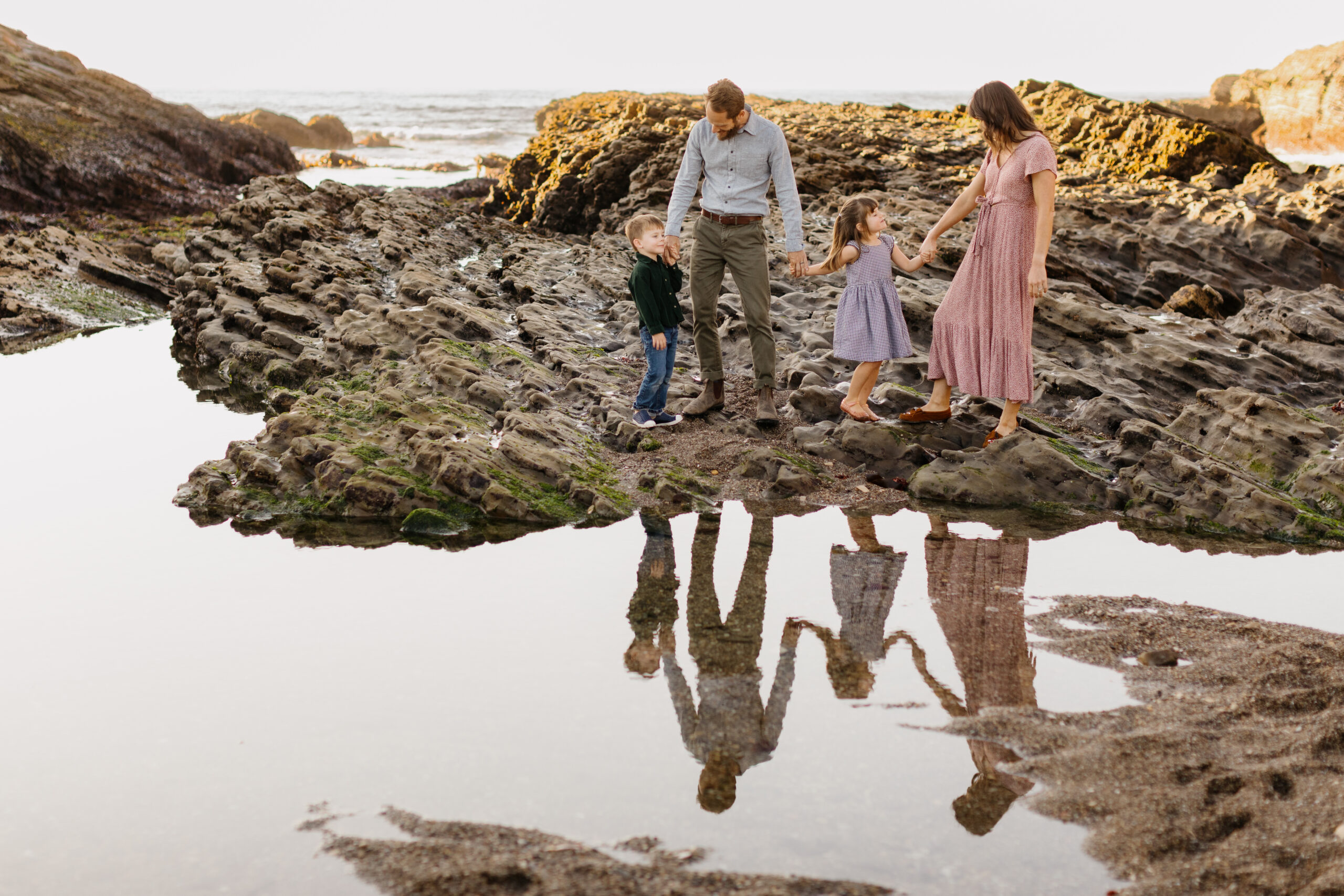 Parents and their two small children standing on a beach rock in Los Osos, ca at Montana De Oro state Beach, Their reflection shows in a tidpool in front of them at their family photo session by family photographer tayler enerle