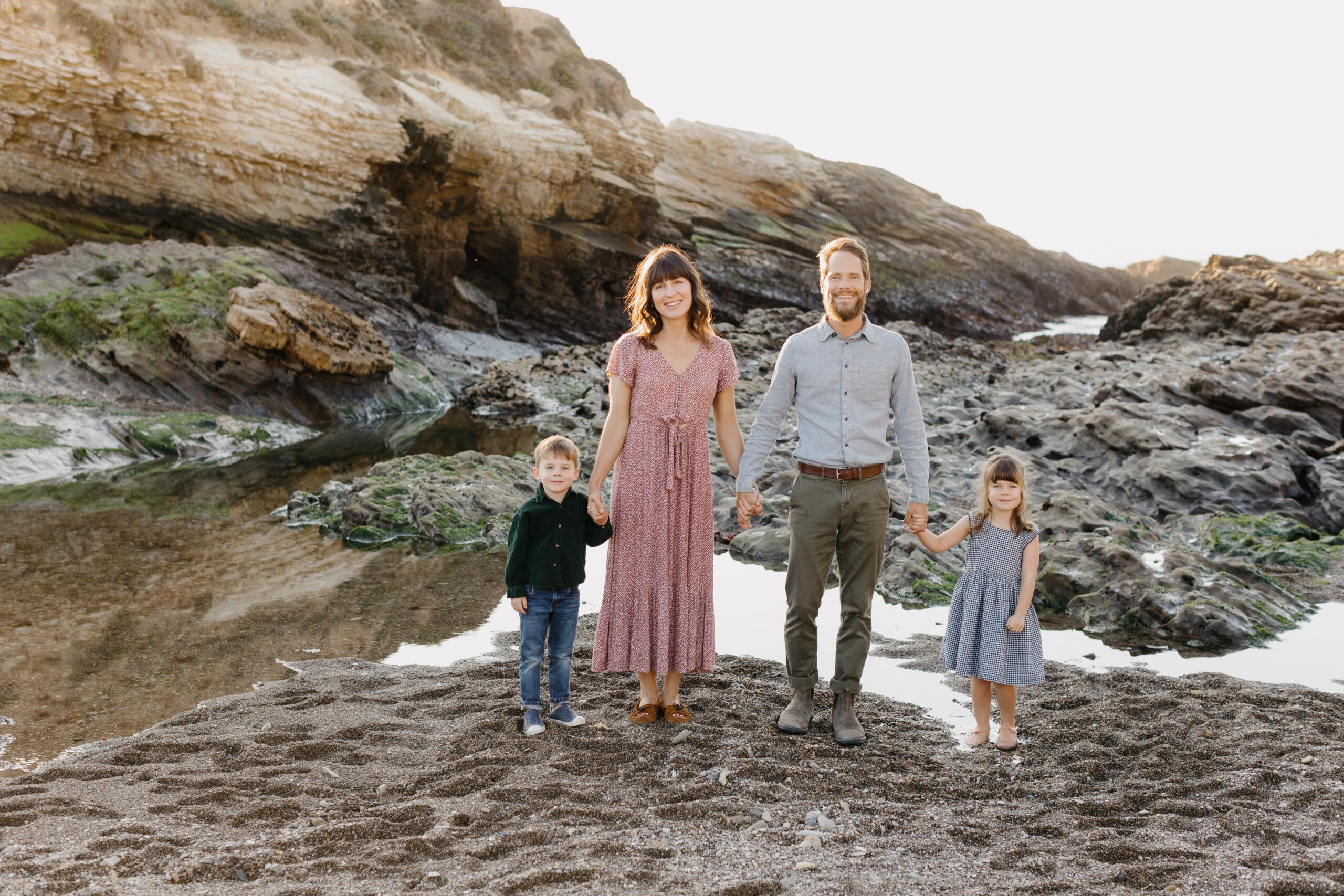 Family holding hands on the sand. in front of rocks at the beach and smiling at the Montana De Oro Family photographer Tayler Enerle's camera