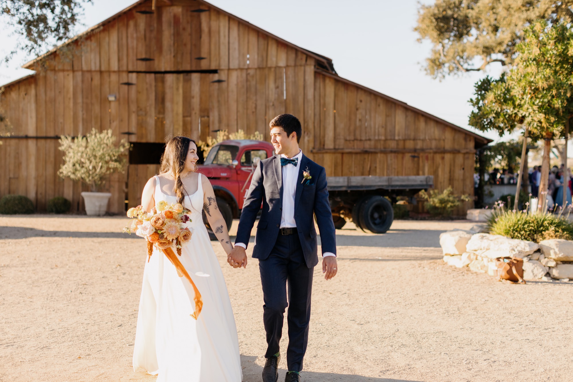 couple walking hand in hand and smiling at eachother at their 1800 El Pomar wedding in templeton, california in front of large brown barn and vintage red truck with el pomar written on it by san luis obispo photographer, tayler enerle