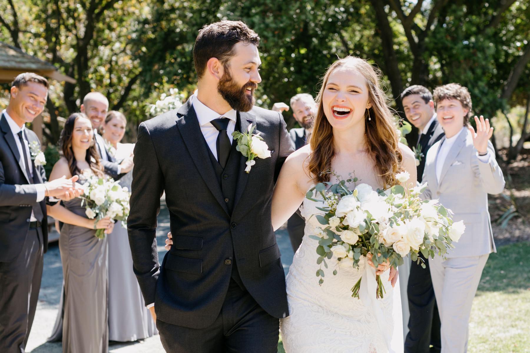 Bride and groom laughing and smiling at each other with their wedding party behind them at their wedding ceremony at cal poly arboretum, and their reception at the penny in san luis obispo california