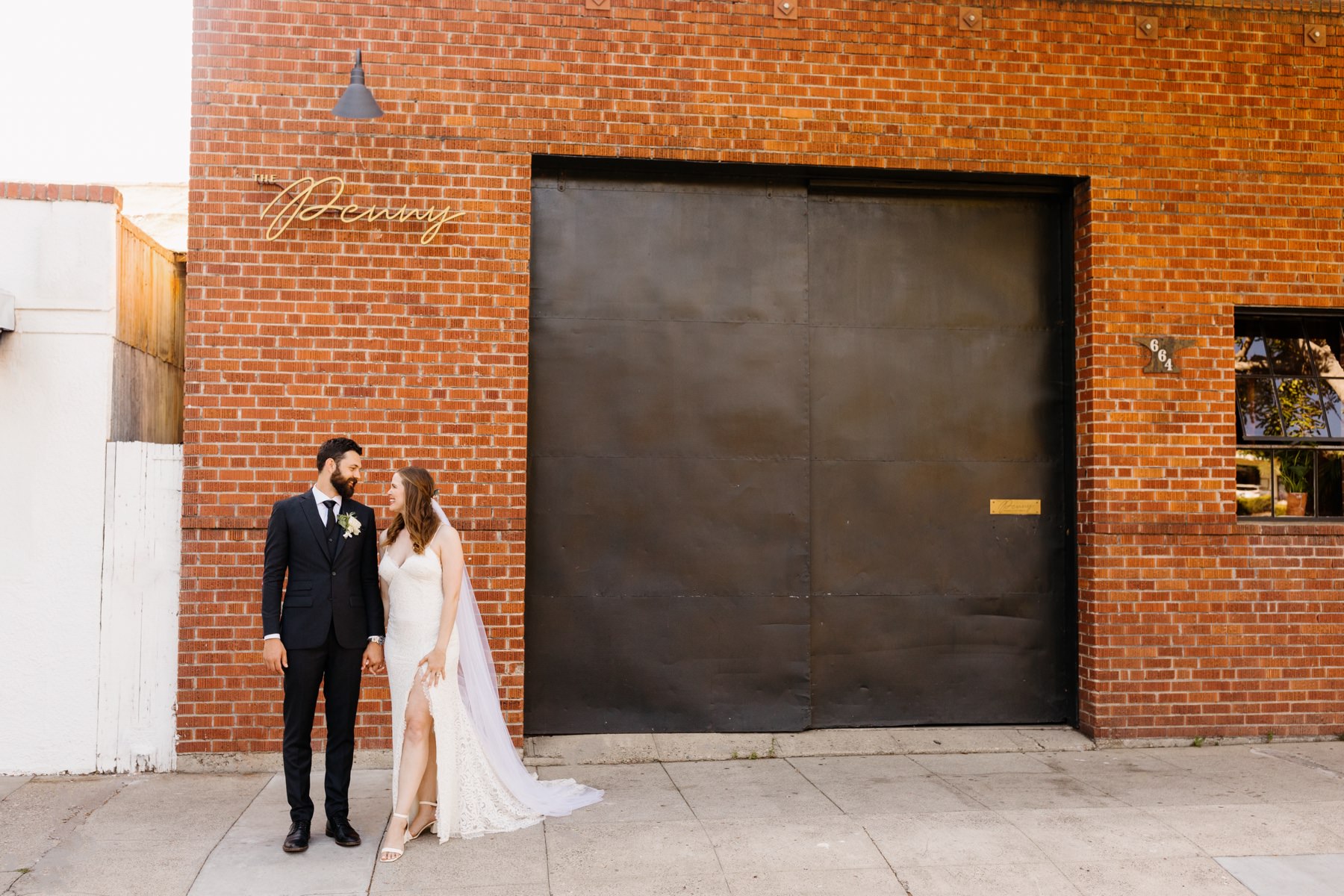 a newlywed couple standing outside of the penny where their wedding reception is being held in san luis obispo, california, couple is looking at eachother in front of the brick wall and black garage door