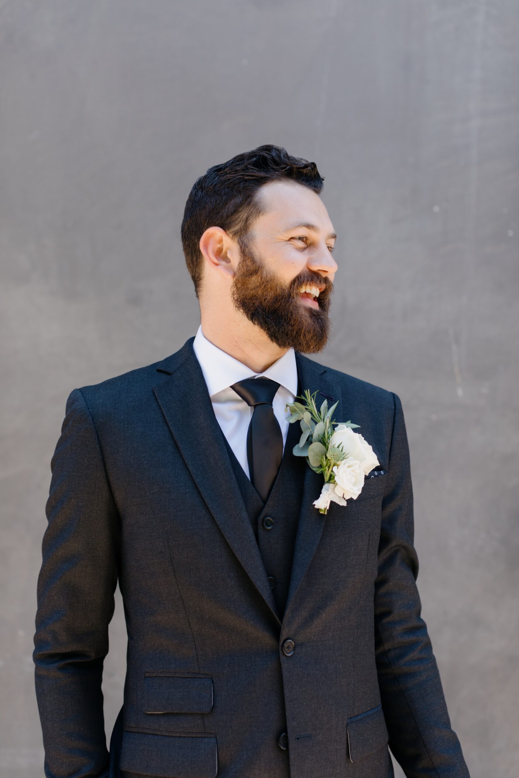 A groom in a black suit with white shirt and black tie with a white flower boutonniere, smiling from a side profile for a portrait again a gray wall on his wedding day at the Penny in San Luis Obispo california