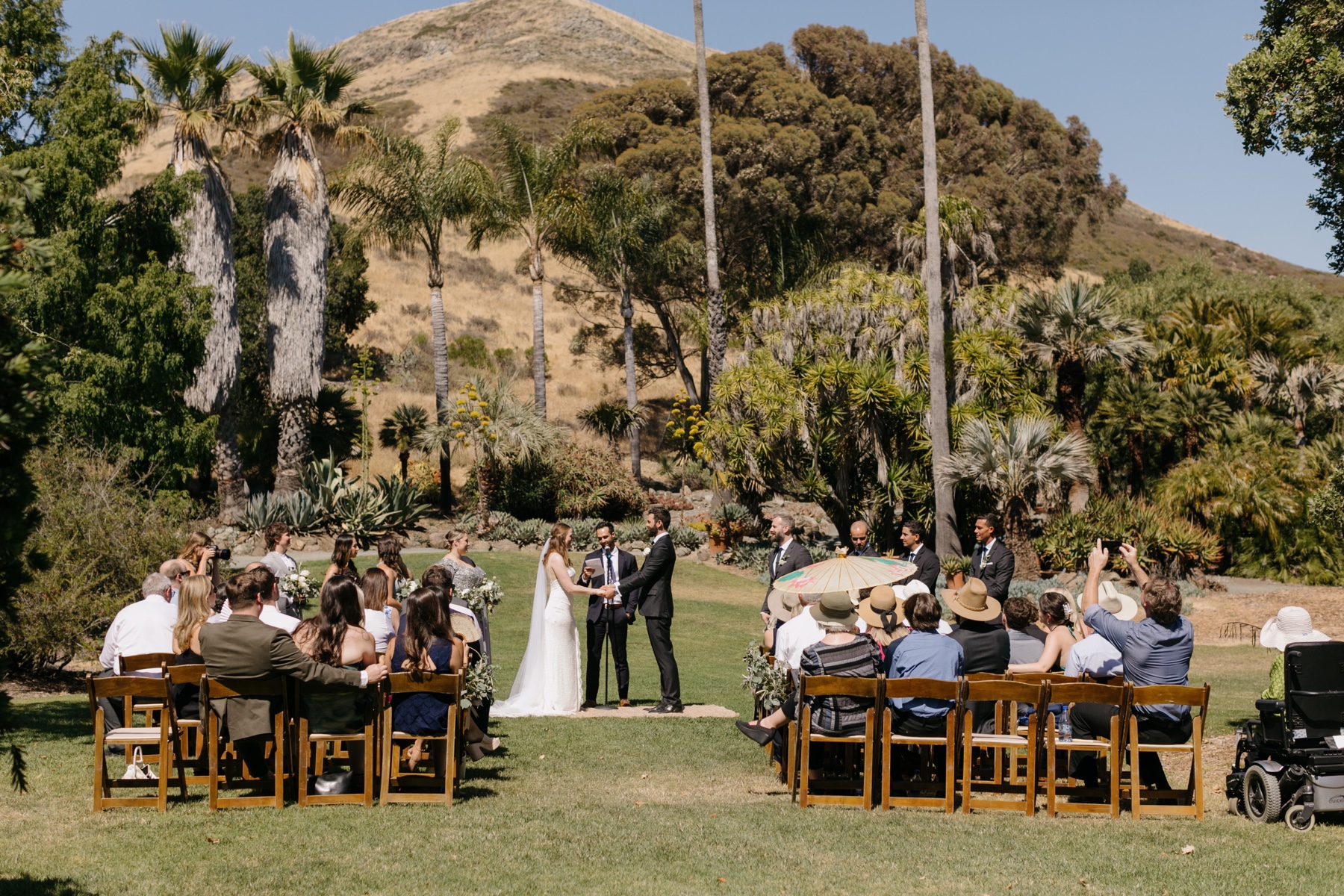 Wide photo of a wedding ceremony at the cal poly arboretum with the hills of slo and trees in the background, slo graduates have their wedding ceremony 