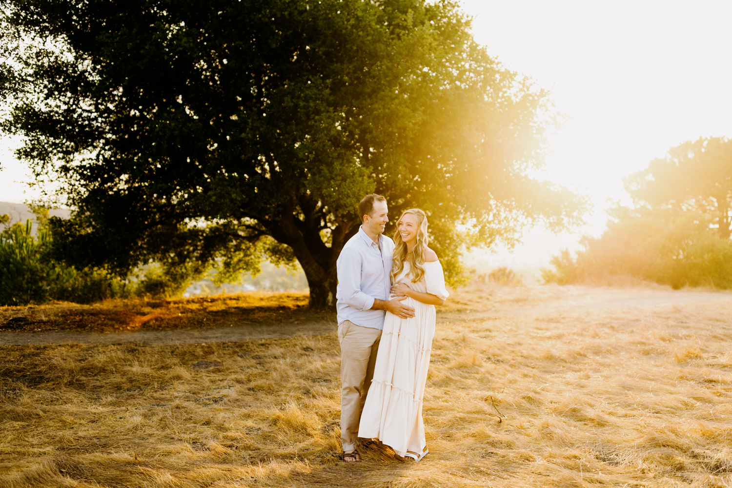 partner embracing his pregnant wife and laughing together at sunny maternity session in San Luis Obispo, ca