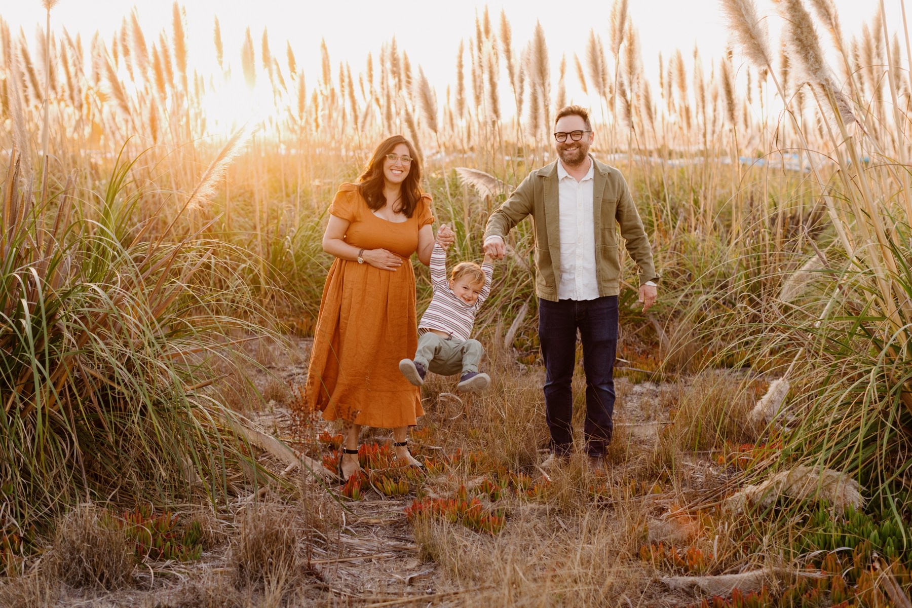 A pregnant mother, a father and their young child swinging the child by the hands and playing together at their los osos maternity session at Cuesta inlet in Los Osos California. They are standing on iceplant in front of pampas grass at sunset overlooking the cuesta inlet Bay