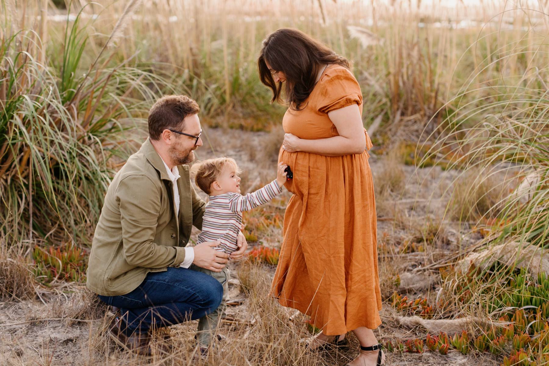 A small child holding his toy car to his mom's pregnant belly playing with little sister while dad holds him from behind. They are standing on wild grass and iceplant on the Cuesta inlet bay in Los Osos California with pampas grass behind them for their sunset maternity session with Tayler Enerle Photography