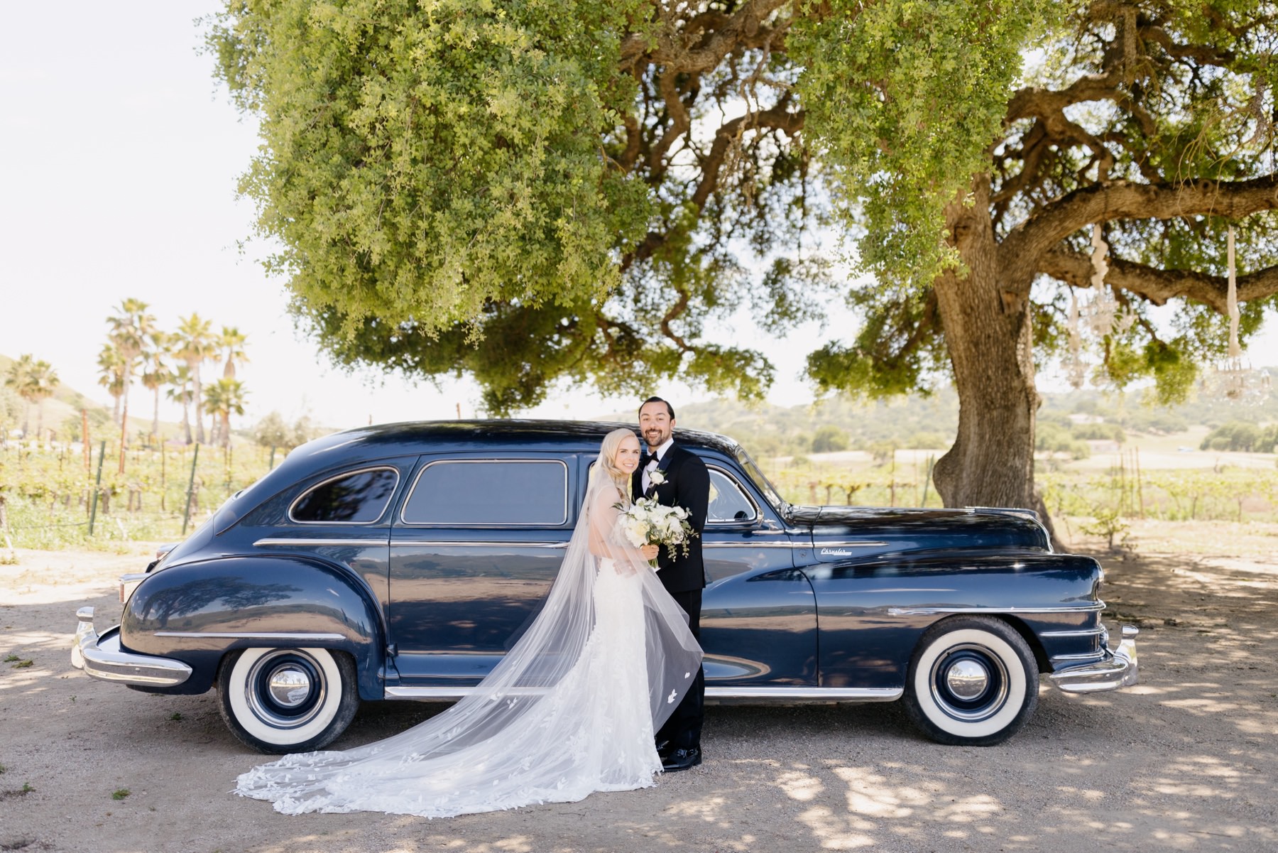 Horizontal image of Bride and groom embracing and smiling with foreheads touching standing in front of an oak tree and a vintage car on their wedding day at Cass Winery in Paso RObles california photographed by Tayler Enerle 