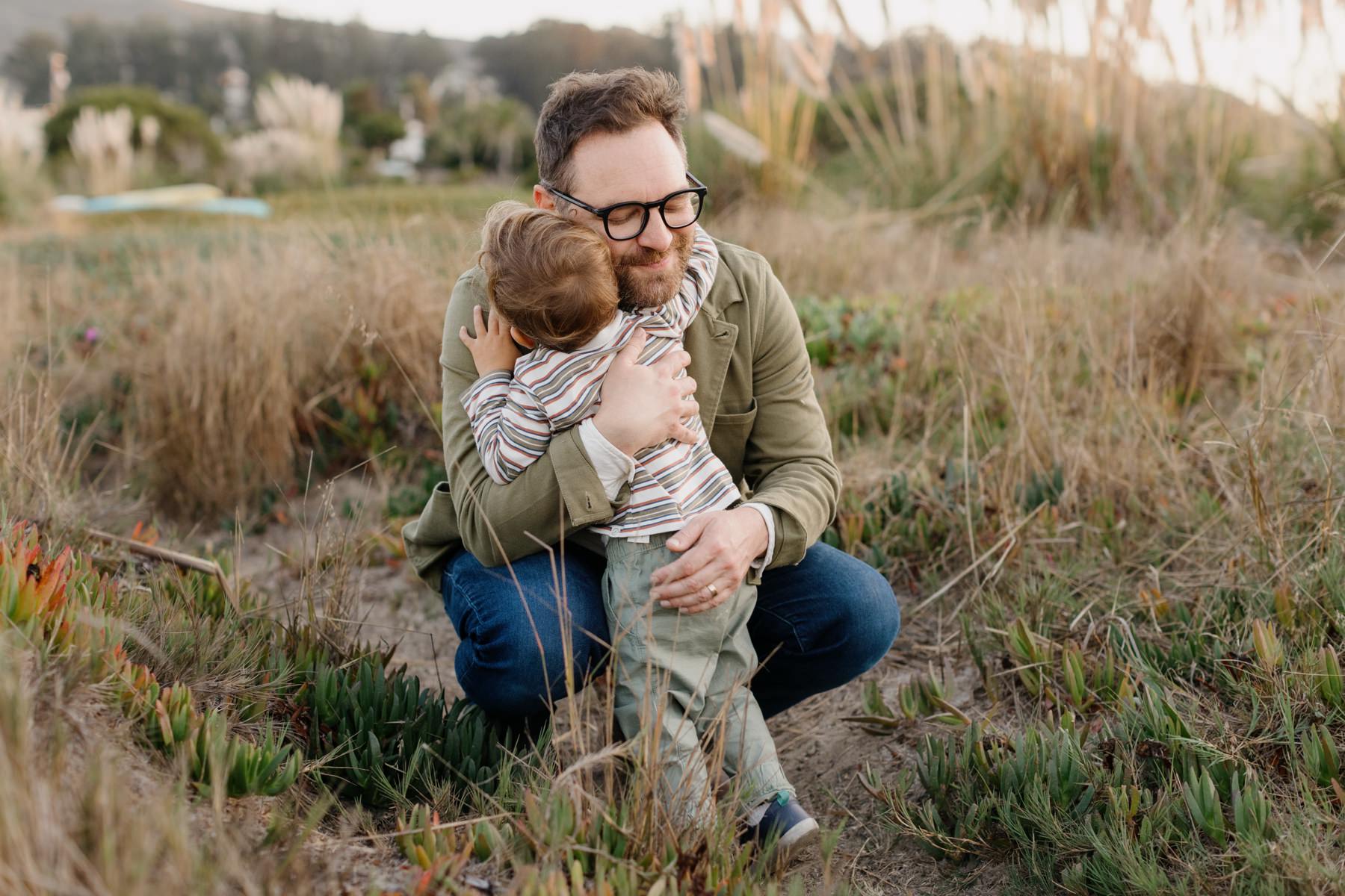 Sweet image of a young boy hugging his father as the father kneels down on the iceplant and wild grass they are on at the Cuesta Inlet with Pampas grass in the background in Los osos California at their maternity session by tayler enerle photography