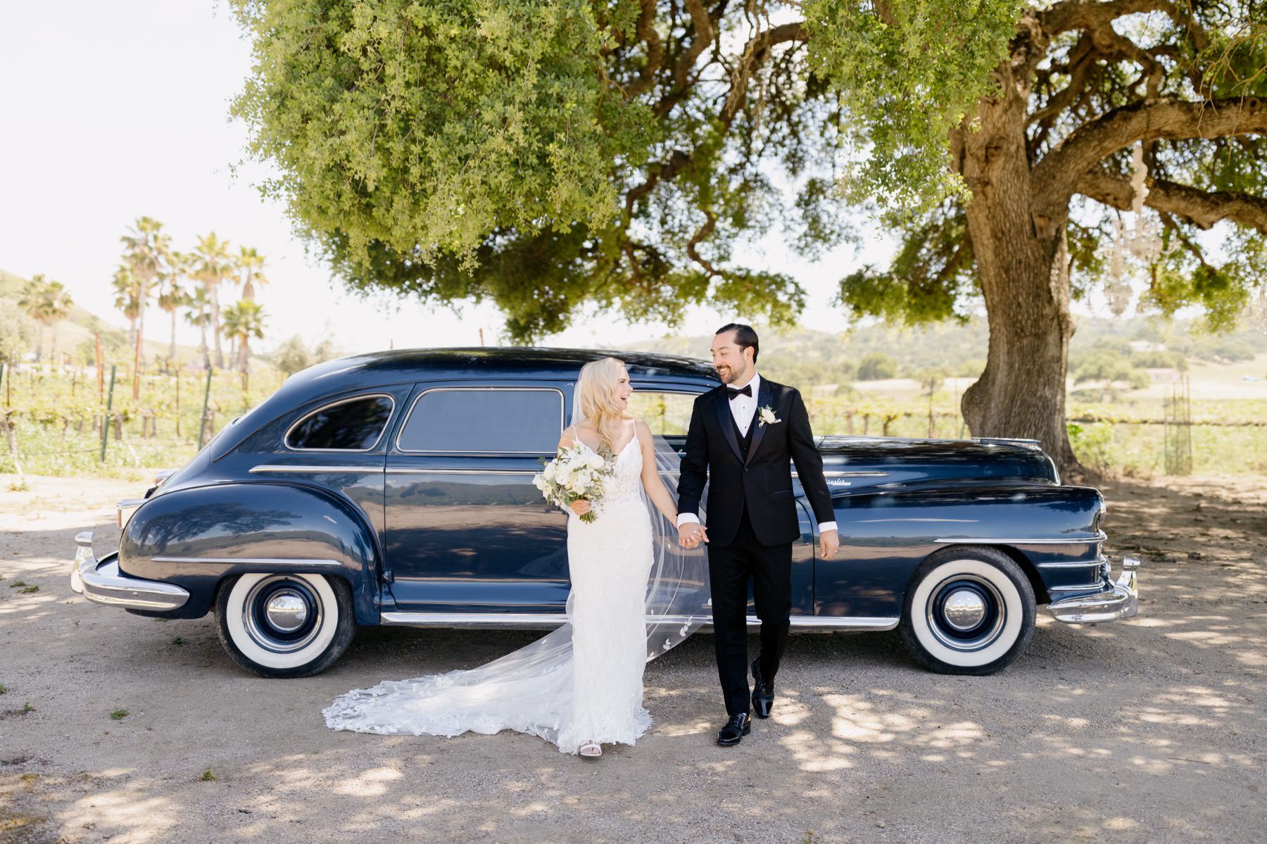 Horizontal image of Bride and groom holding hands and smiling at each other walking towards the camera and standing in front of an oak tree and a vintage car on their wedding day at Cass Winery in Paso RObles california photographed by Tayler Enerle