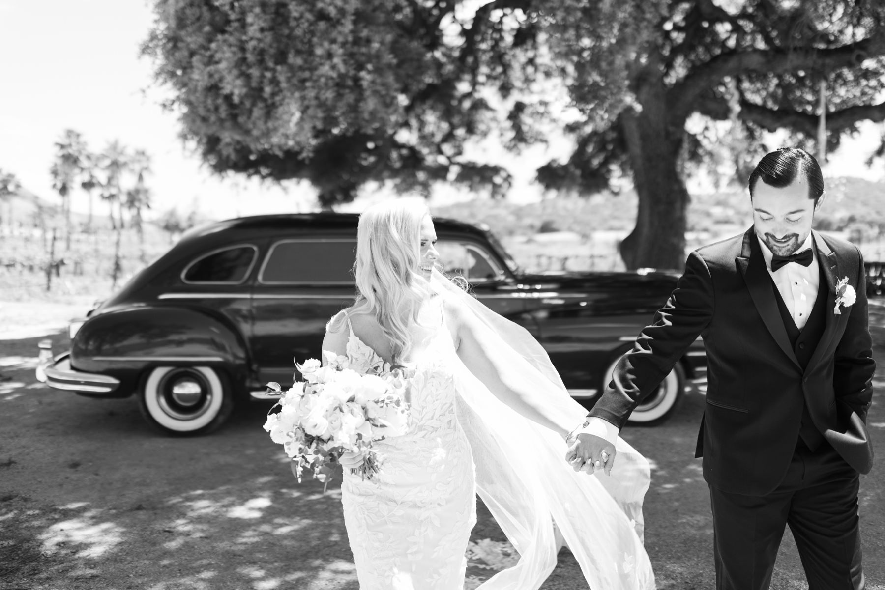 Black and white image of Bride and groom walking towards camera holding hands and smiling at eachother while the bride's veil gets blown behind her in the wind during their portrait session on the fun Cass Winery wedding day photographed by tayler enerle Photography