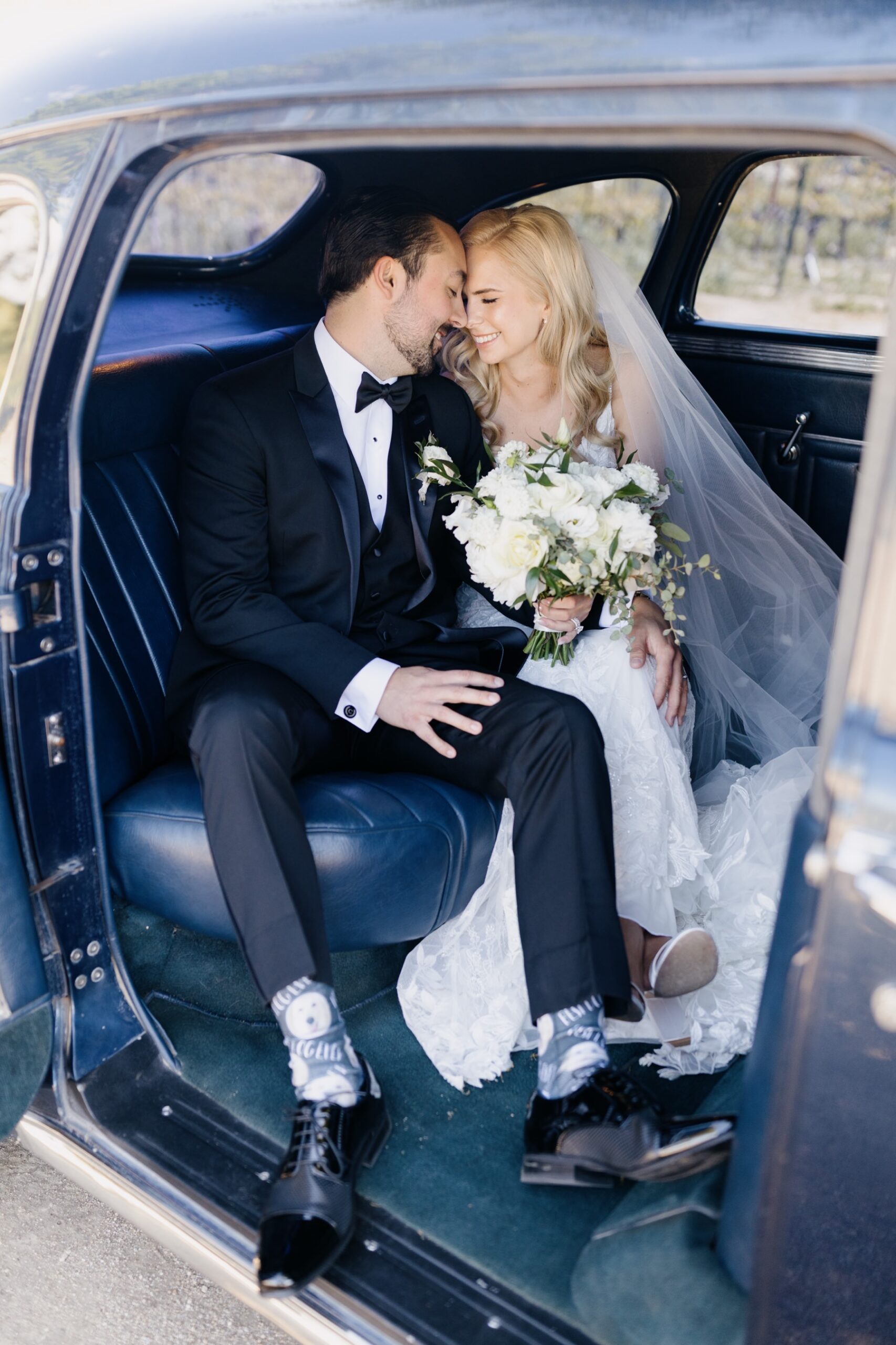 vertical image of Newly married couple sitting face to face and smiling in their vintage car after their ceremony and before their fun Cass Winery wedding in Paso Robles California during their portraits session with Tayler Enerle photography