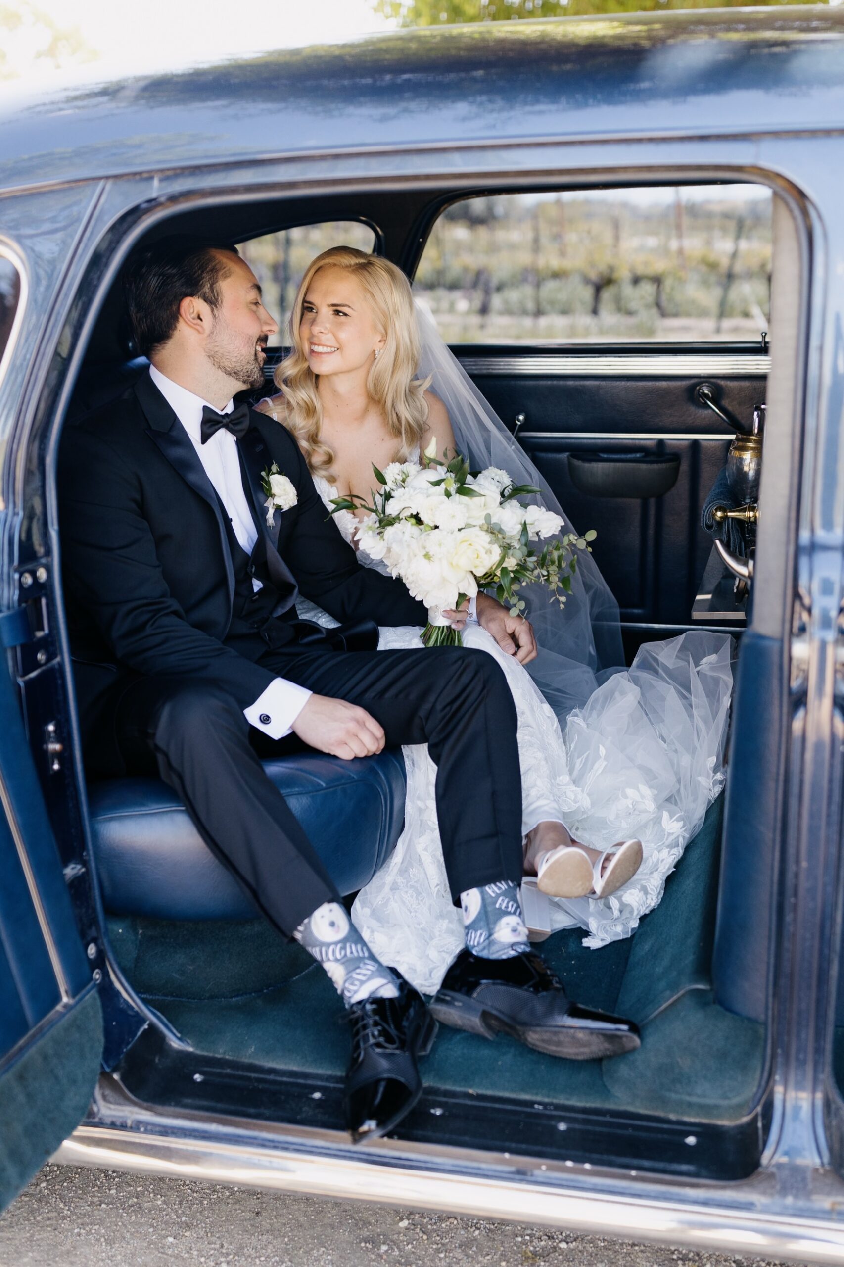 vertical image of Newly married couple sitting face to face and smiling in their vintage car after their ceremony and before their fun Cass Winery wedding in Paso Robles California during their portraits session with Tayler Enerle photography