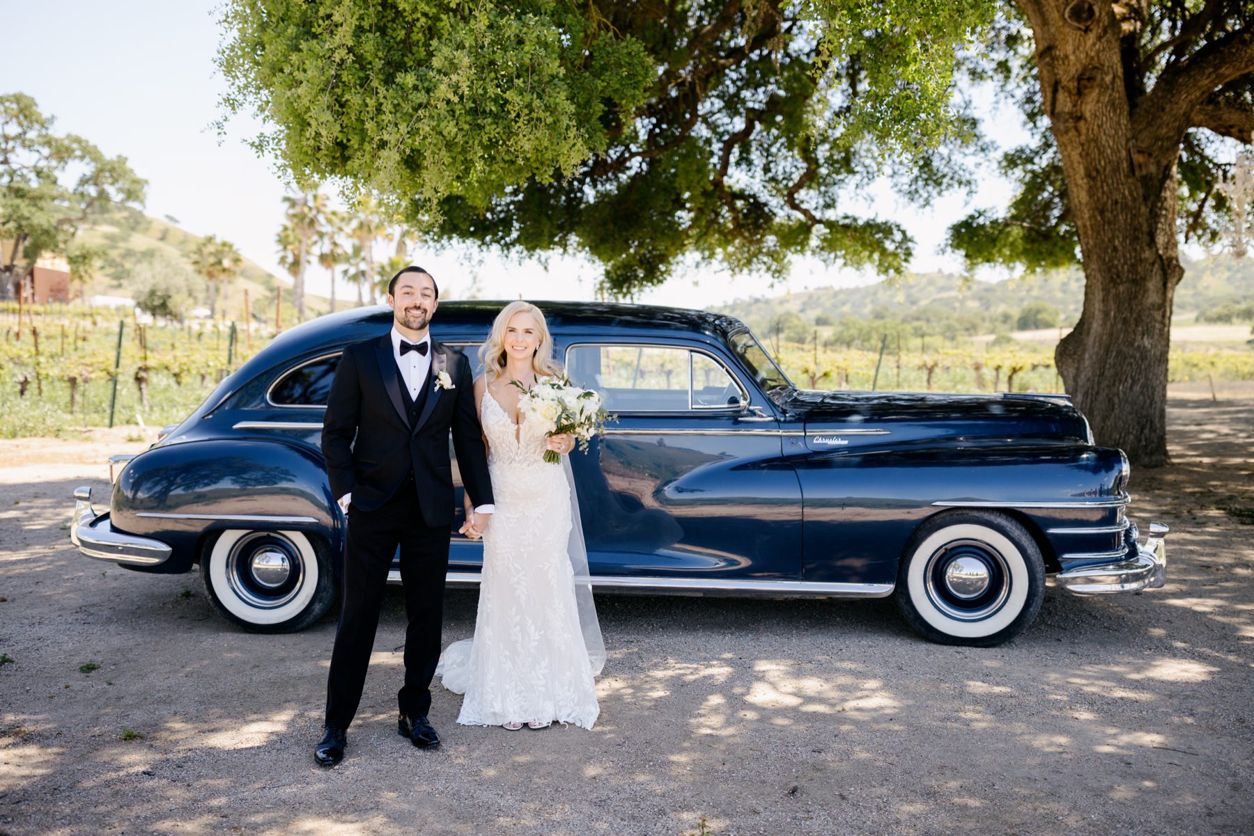 newly married couple standing in front of a vintage car holding hands and smiling at the camera under and oak tree at their portrait session at Cass Winery in Paso Robles California taken by Tayler Enerle Photography 